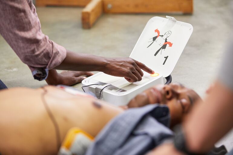 Bewusstloser Mann liegt am Boden, ein AED ist an angeschlossen und wird von einem Helfer bedient.