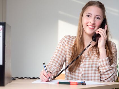 Azubine sitz im Büro und telefoniert. Sie hat einen Kuli in der Hand und macht sich Notizen