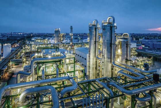 Eine moderne Industrieanlage mit großen Rohren und Türmen bei Dämmerung, beleuchtet von zahlreichen Lichtern.