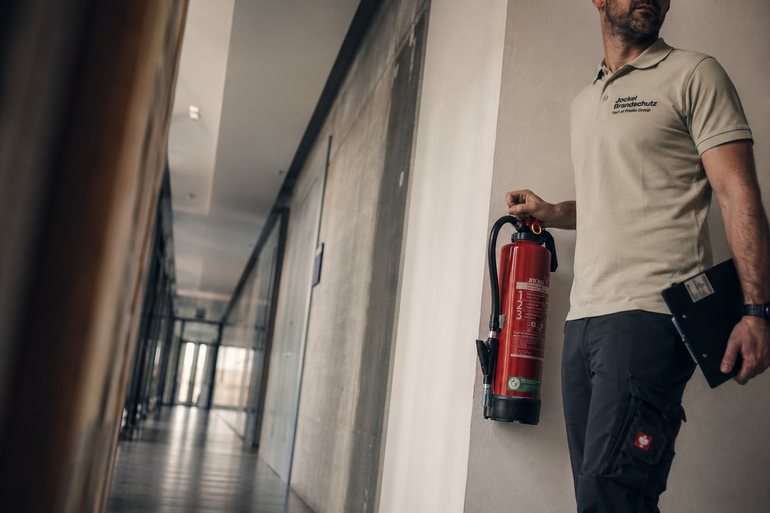 Ein Techniker steht in einem modernen Flur und hält einen Feuerlöscher sowie ein Notizbuch in der Hand.